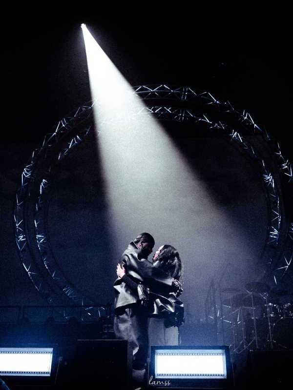 Two performers interacting on stage, highlighted by a single intense beam of light in a dark venue. — concert photography London by Lamss Media